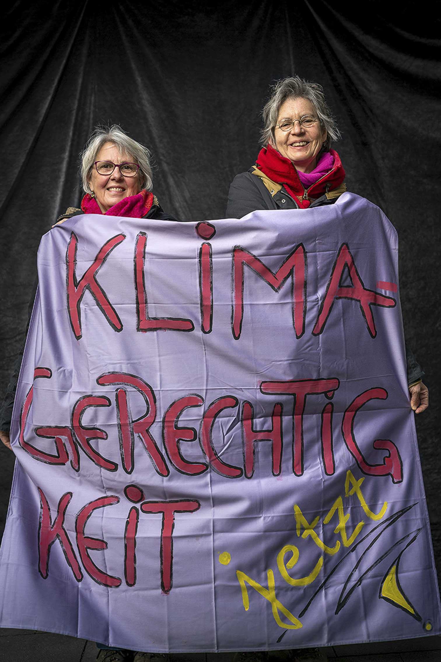 Zwei Frauen mit Banner für Klimagerechtigkeit vor Studiohintergrund auf einer FfF Demo, lächelnd.